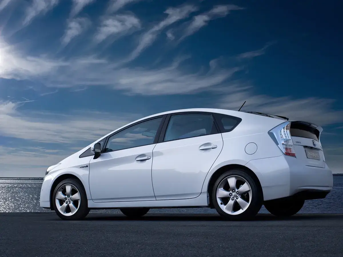 Biały Toyota Prius zaparkowany przy wodzie pod błękitnym niebem z chmurami.