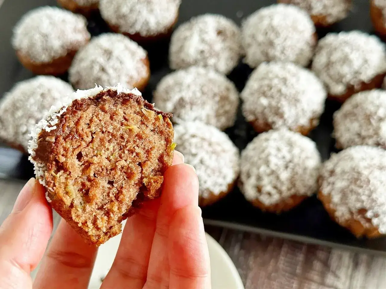 Pyszne muffinki z cukinią, oblane czekoladą i posypane wiórkami kokosowymi. Jedna muffinka jest przekrojona, ukazując wilgotne wnętrze.