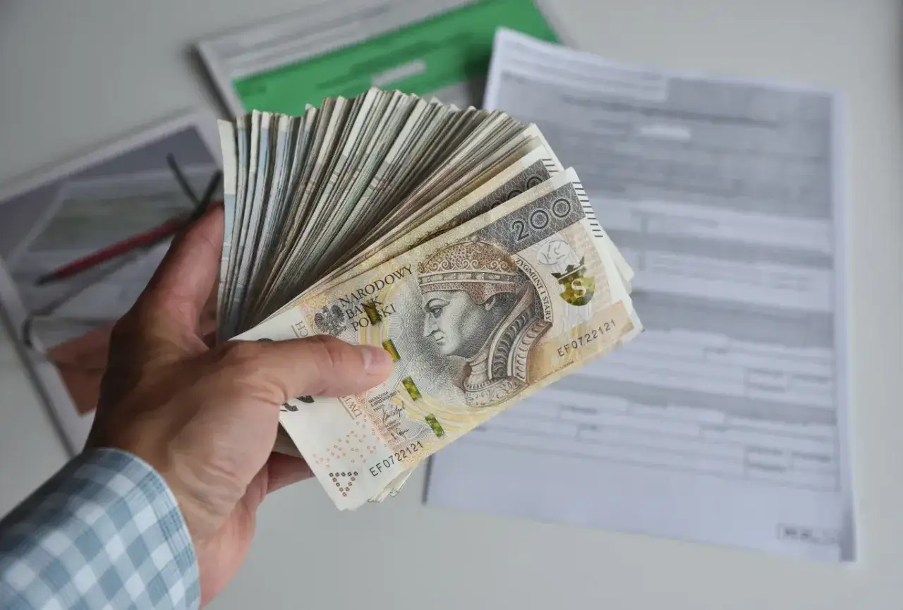 Dłoń trzyma plik banknotów 200 zł. W tle dokumenty, być może dotyczące tego, ile godzin to 0.5 etatu.