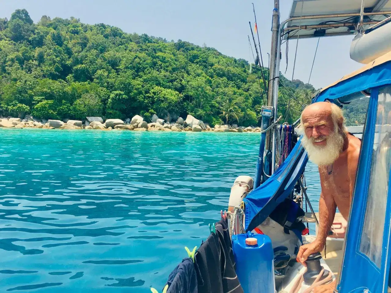 Gangerl Clemens genießt die Fahrt auf seinem Segelboot vor einer üppig grünen Insel mit türkisfarbenem Wasser.