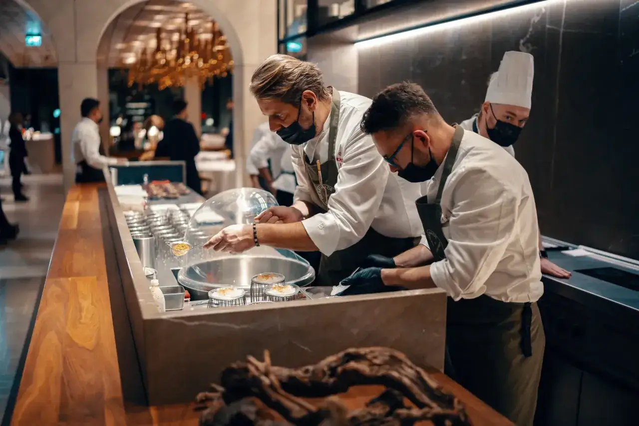 Szefowie kuchni w atelier amaro przygotowują dania. W tle widać eleganckie wnętrze restauracji.