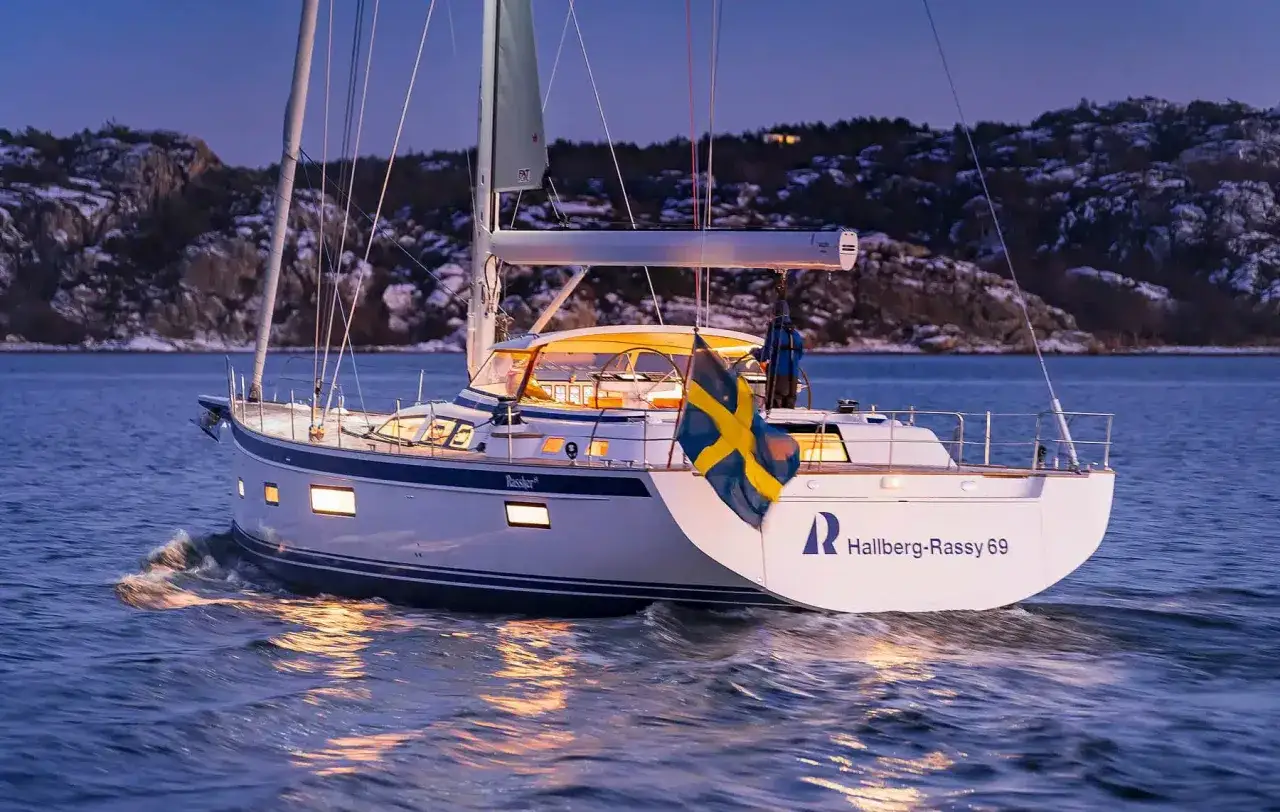 Eine Hallberg-Rassy 69 gleitet bei Dämmerung über das Wasser, die schwedische Flagge weht im Wind.