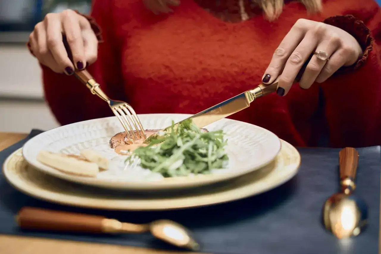 Une femme savoure un plat, couteau et fourchette dorés à la main. L'élégance de la table évoque l'art de la table France.