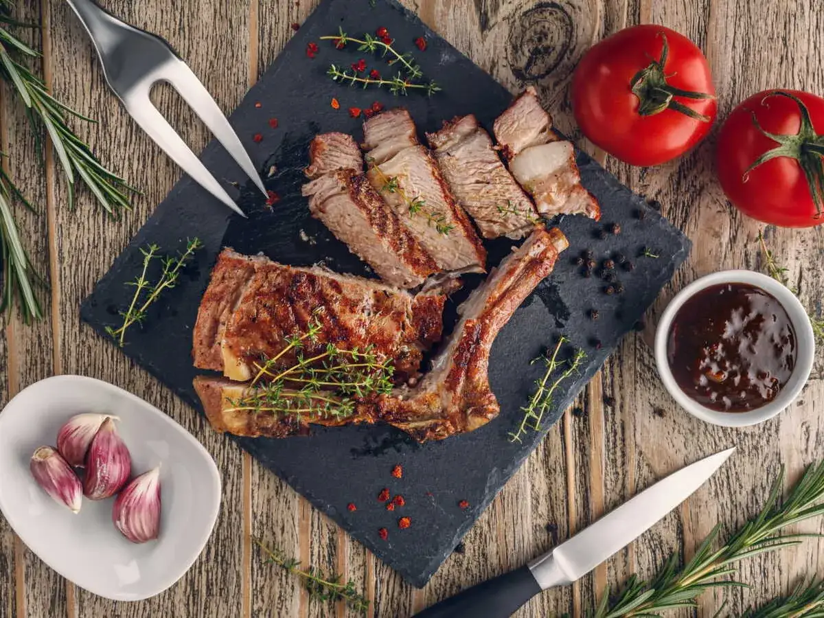 Côte de veau grillée tranchée sur une planche d'ardoise, garnie d'herbes, avec des tomates, de l'ail et une sauce.