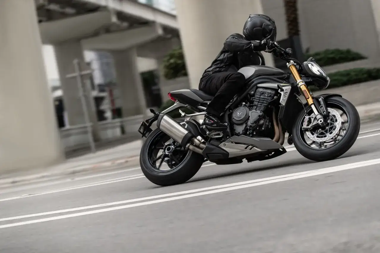 Motocyklista w kasku i skórzanej kurtce pochyla się na swoim motocyklu Triumph Speed Triple, pokonując zakręt na miejskiej drodze.