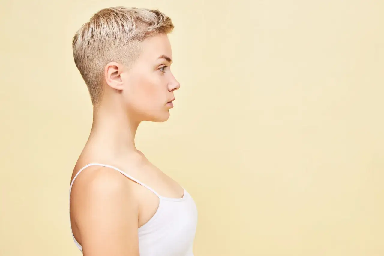 Perfil de una mujer joven con un moderno corte pixie rubio, luciendo un top blanco.