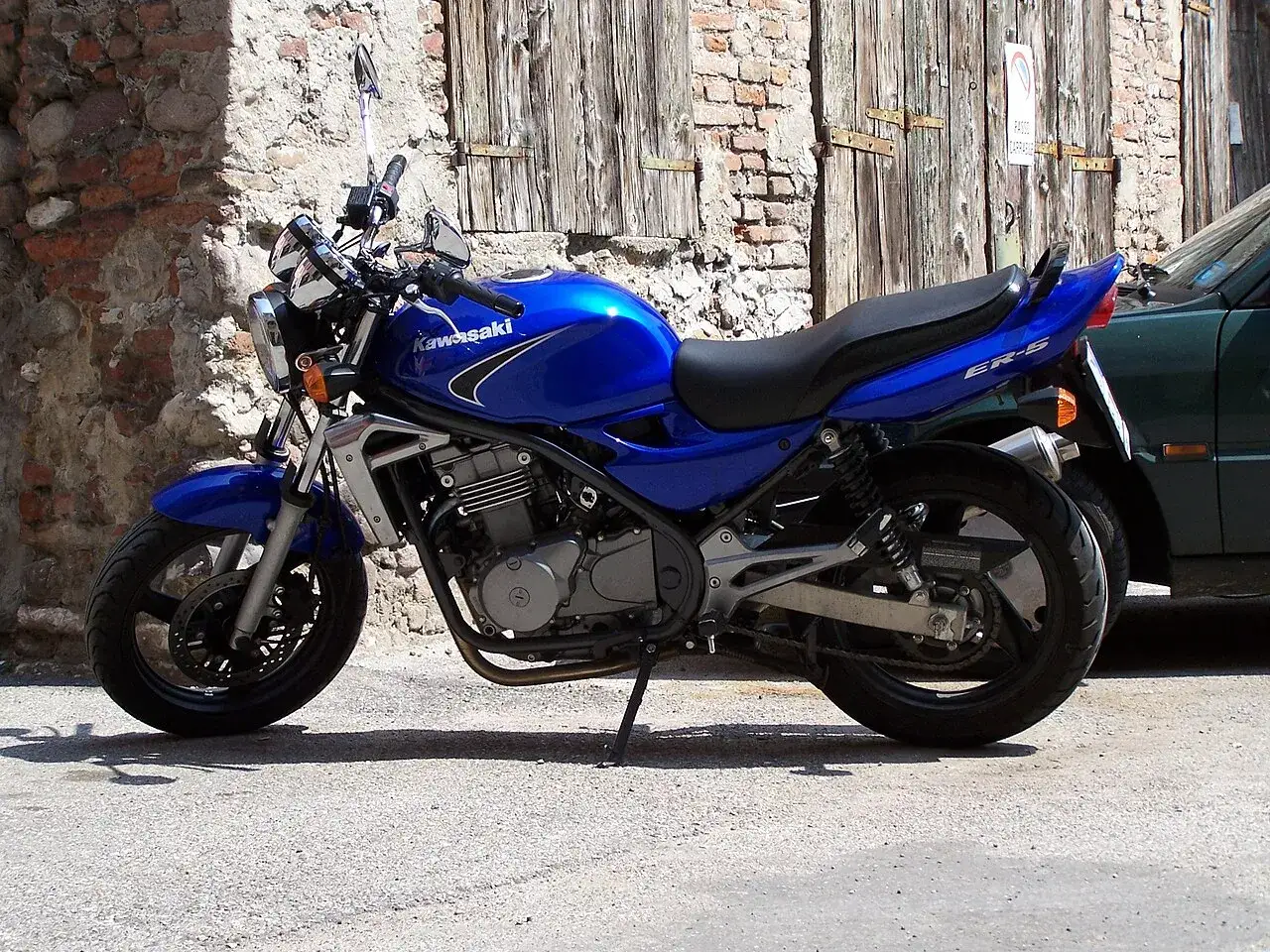 Błękitny motocykl Kawasaki ER-5 zaparkowany przy kamiennym murze i drewnianej bramie.