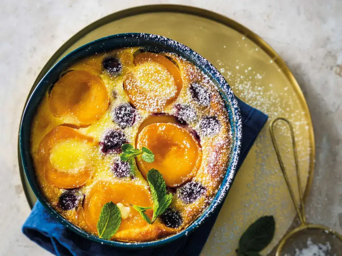 Clafoutis abricot sans gluten, doré et saupoudré de sucre glace, garni de fruits frais et de feuilles de menthe.