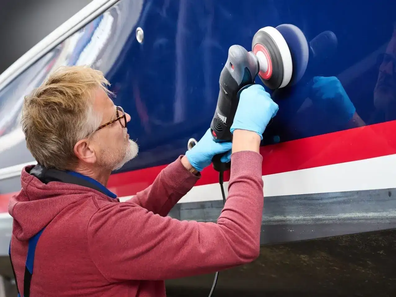 Mann poliert mit Maschine den blauen Bootsrumpf. Das Gelcoat erstrahlt nach dem Polieren wieder in neuem Glanz.