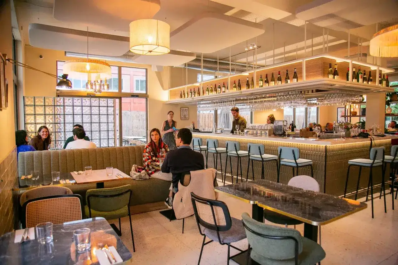 Ambiance chaleureuse d'un restaurant, avec des clients attablés et un bar bien garni. L'esprit du terroir se retrouve dans la sélection des vins et la convivialité.