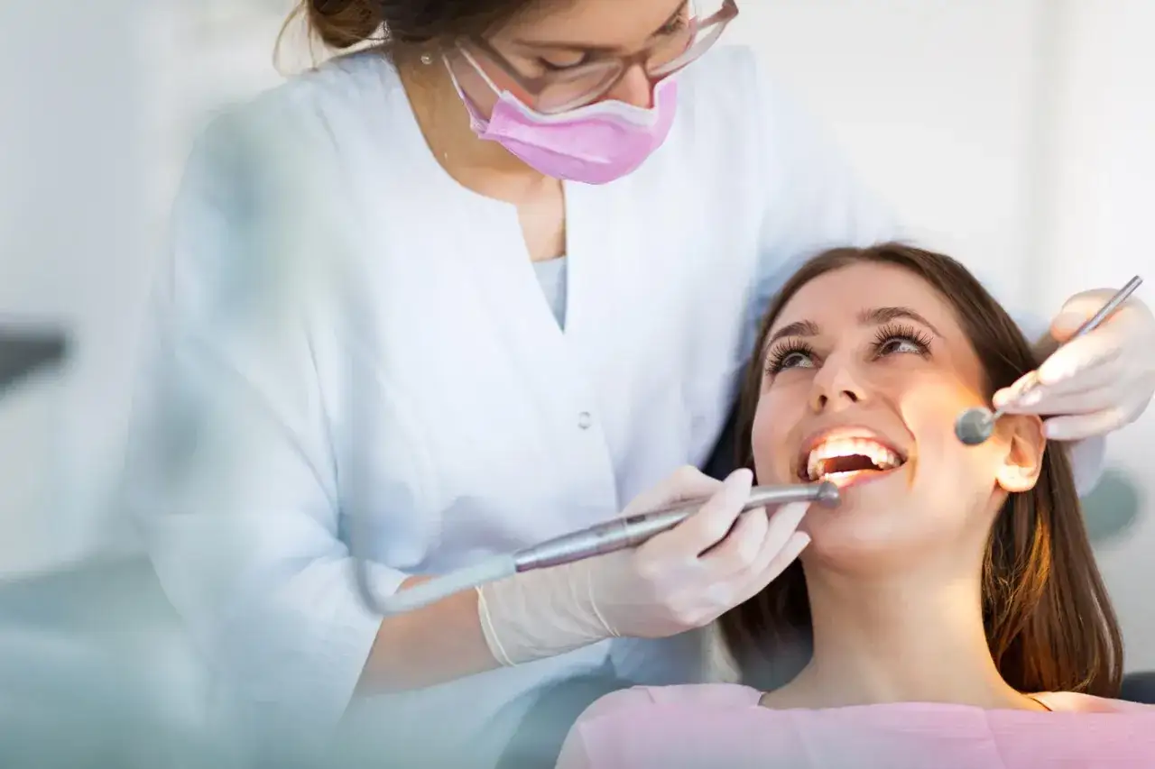 Dentystka w masce i rękawiczkach bada zęby pacjentki za pomocą lusterka i wiertła.