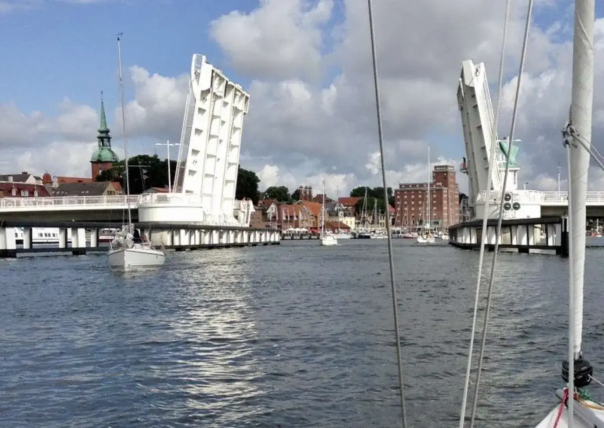 Eine Klappbrücke öffnet sich für Segelboote in Kappeln an der Schlei. Im Hintergrund die Stadt mit Kirchturm und Backsteingebäuden.