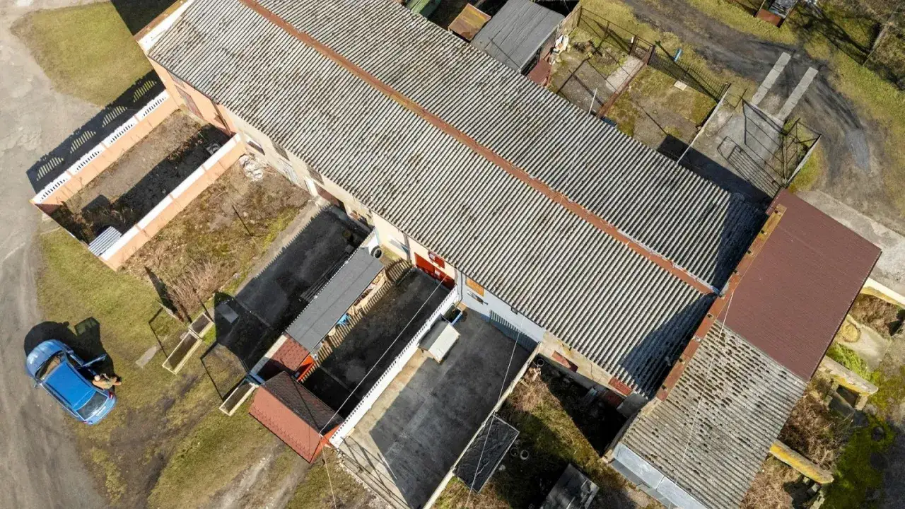 Długi budynek gospodarczy na zgłoszenie z falistym dachem, otoczony innymi mniejszymi obiektami i zielenią. Obok stoi niebieski samochód.