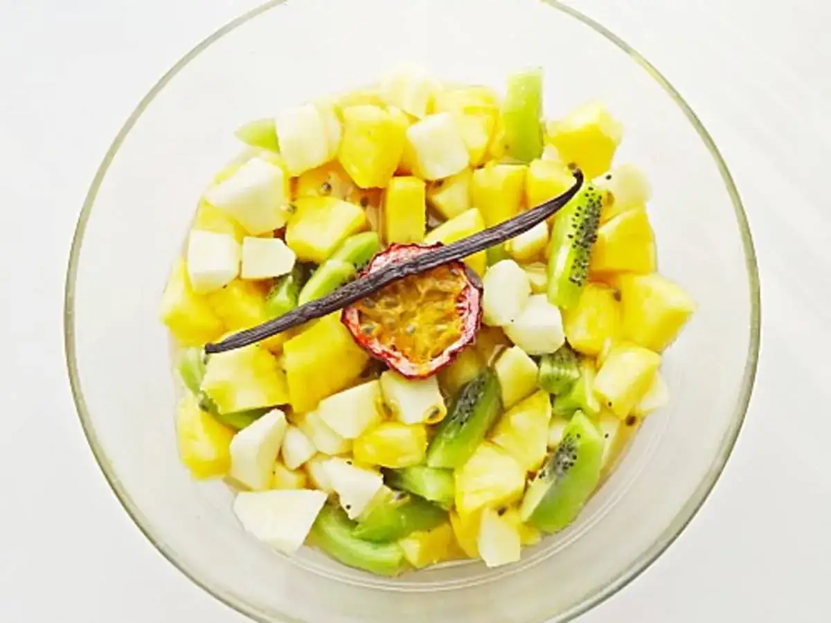 Salade de fruits tropicaux coupés en dés dans un bol en verre, garnie de fruits de la passion et d'une gousse de vanille.