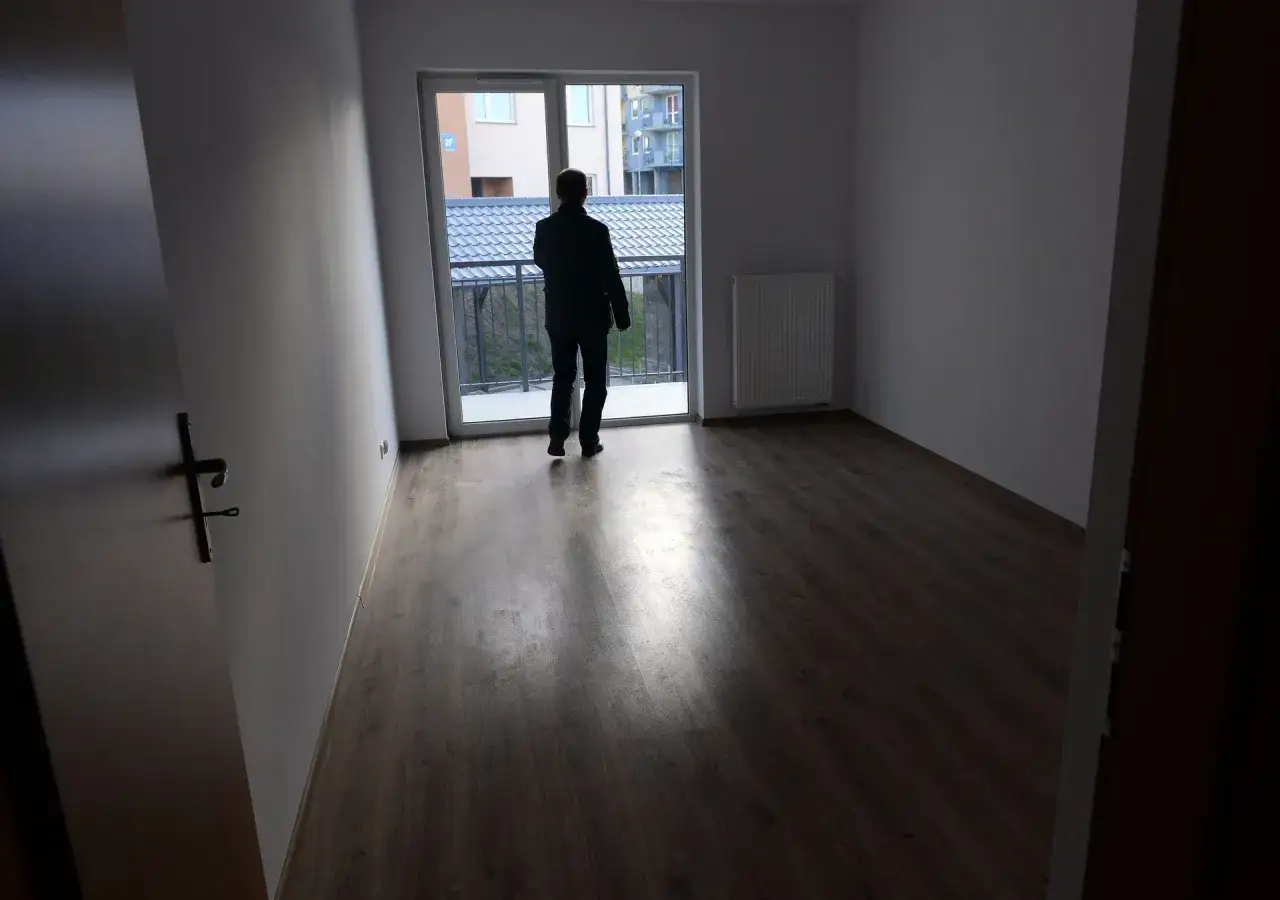 Mężczyzna w pustym mieszkaniu, zastanawia się, gdzie złożyć wniosek o mieszkanie komunalne. Widok na balkon i sąsiednie budynki.