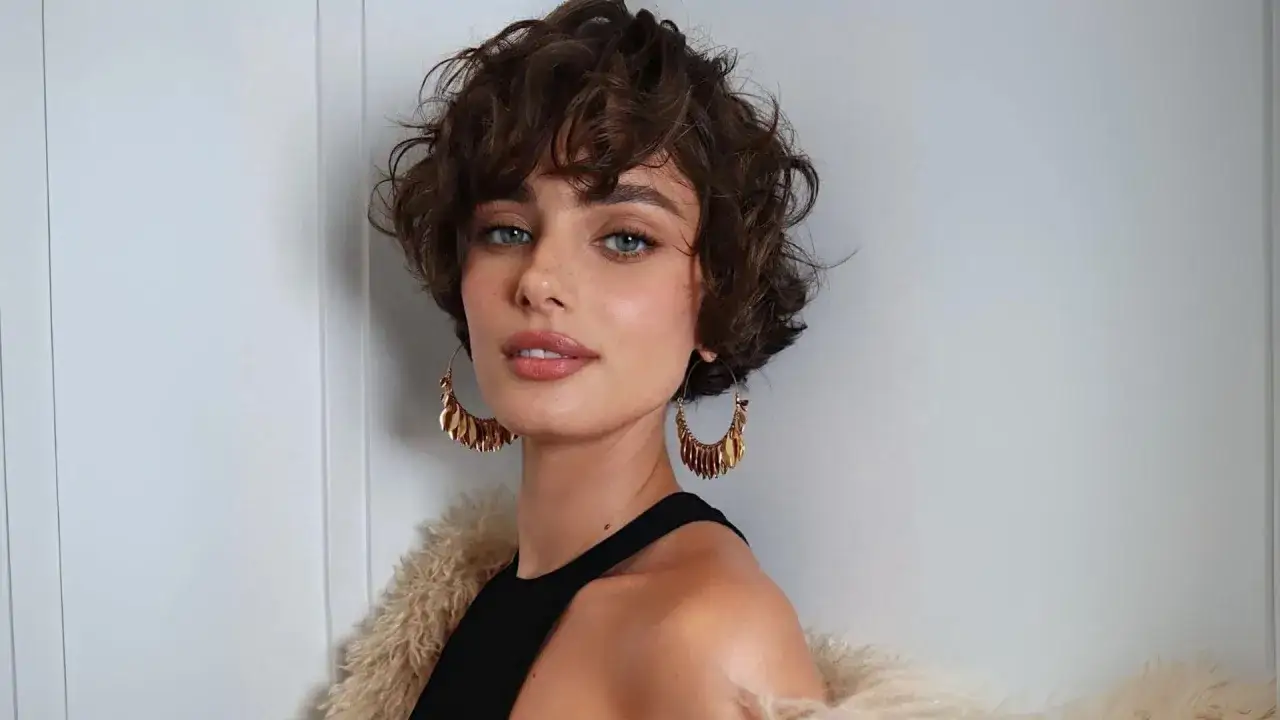 Mujer con un moderno corte shaggy rizado, ojos azules penetrantes y grandes pendientes dorados.