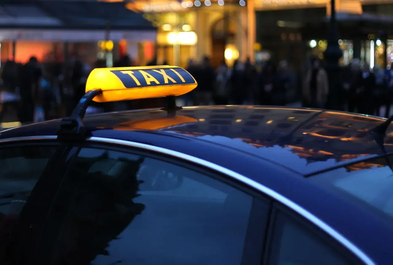 Żółty znak TAXI na dachu ciemnego samochodu, wieczorem, z rozmytym tłem miasta.
