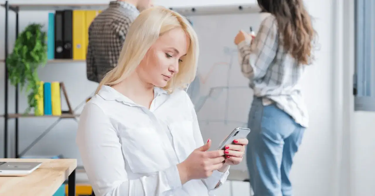 Kobieta z blond włosami korzysta z telefonu, być może używając wyszukiwania głosowego, podczas gdy koledzy pracują przy tablicy.