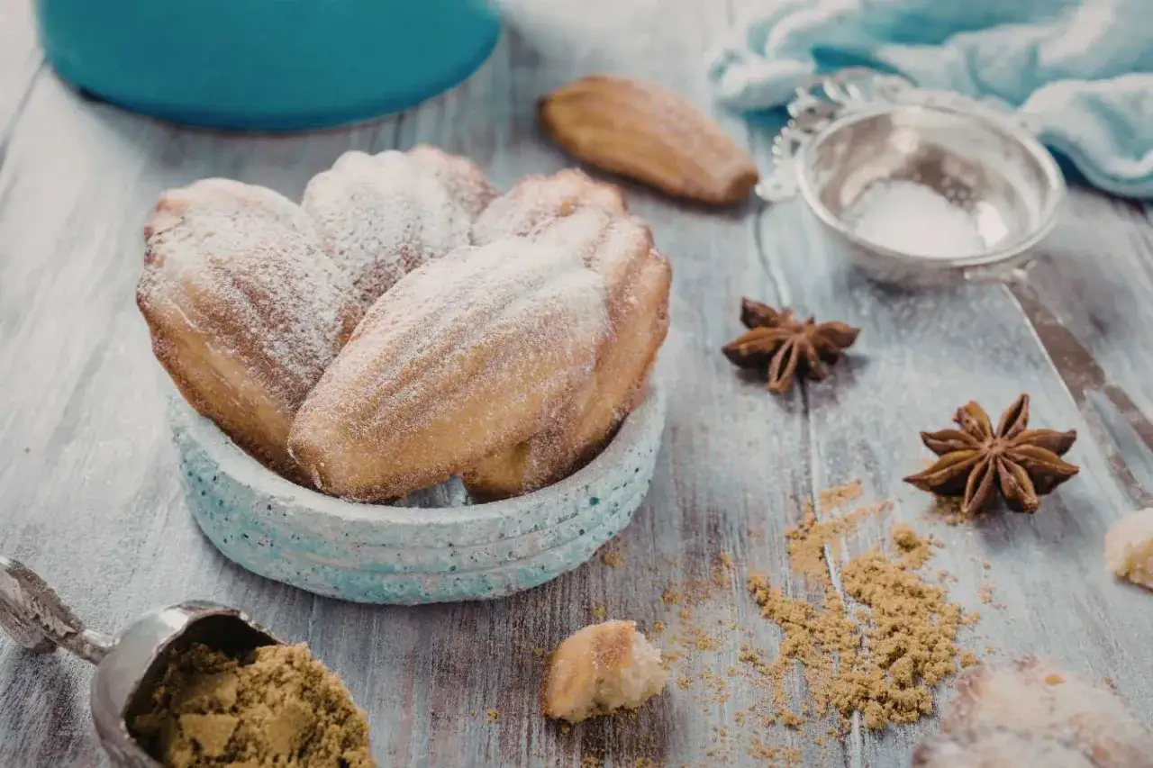 Un bol de biscuits madeleine noisette saupoudrés de sucre glace, avec des étoiles de badiane et une cuillère de poudre sur une table en bois.