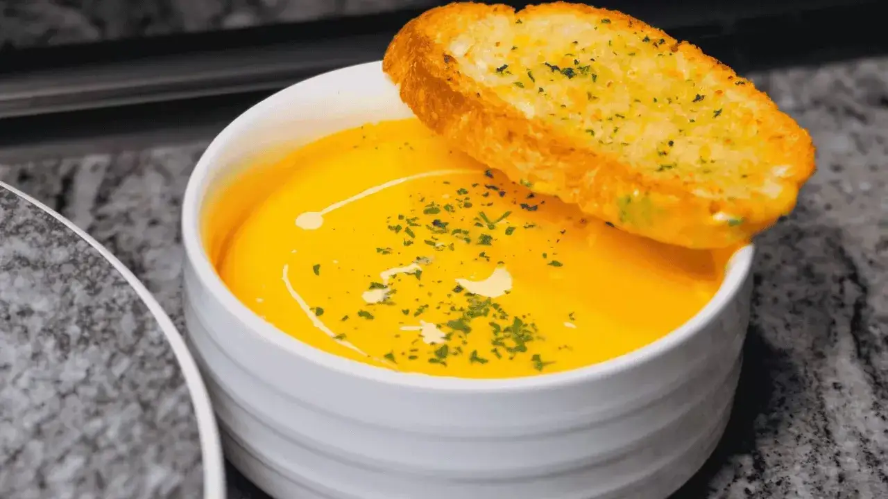 Bol de soupe orange crémeuse garni d'herbes et d'une tranche de pain à l'ail.