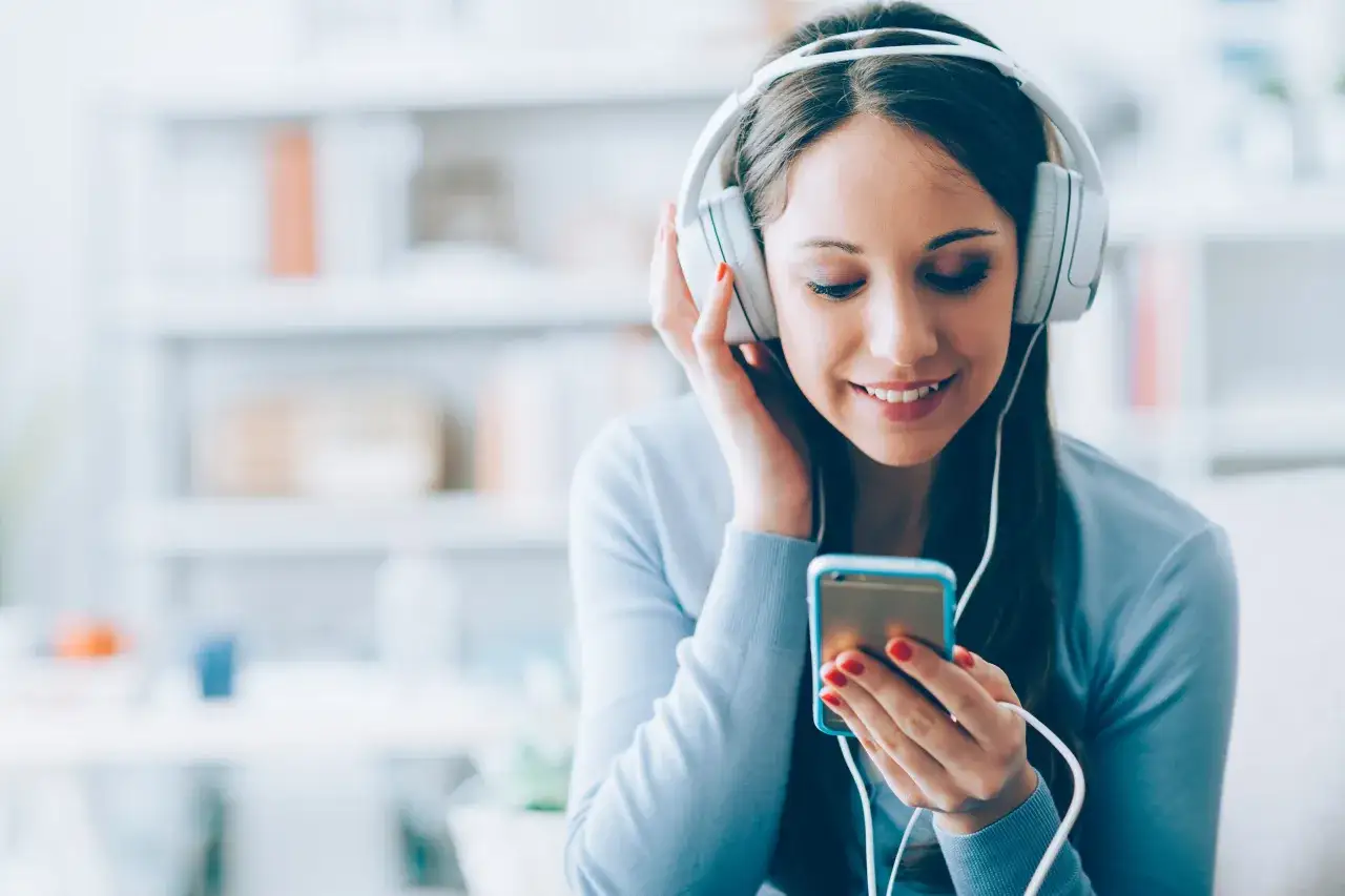 Młoda kobieta w słuchawkach słucha muzyki z telefonu, uśmiecha się.