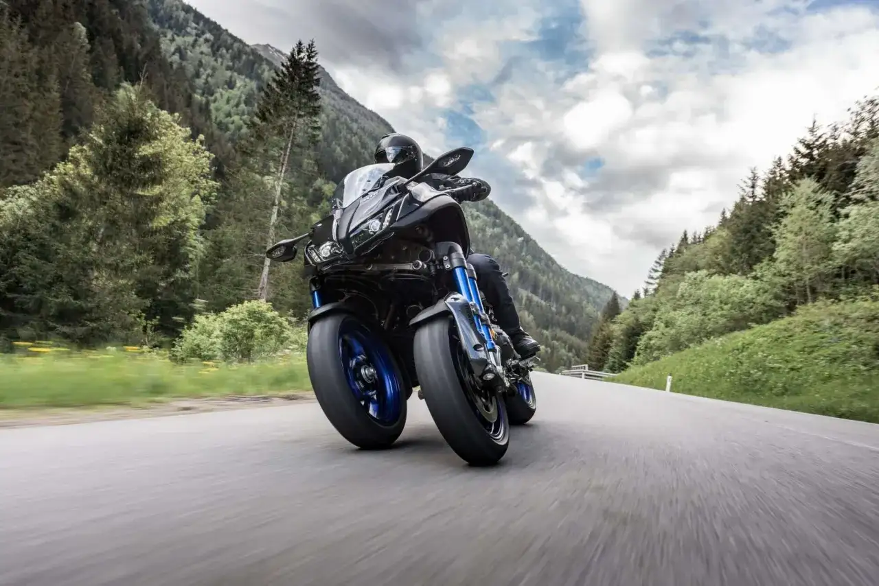 Motocyklista na Yamaha Niken pokonuje zakręt na górskiej drodze, otoczony zielonymi lasami i skalistymi szczytami.