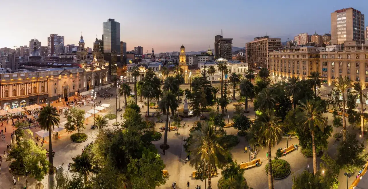 Wieczorny widok na plac w Santiago de Chile, z palmami, budynkami i ludźmi spacerującymi po oświetlonym deptaku.
