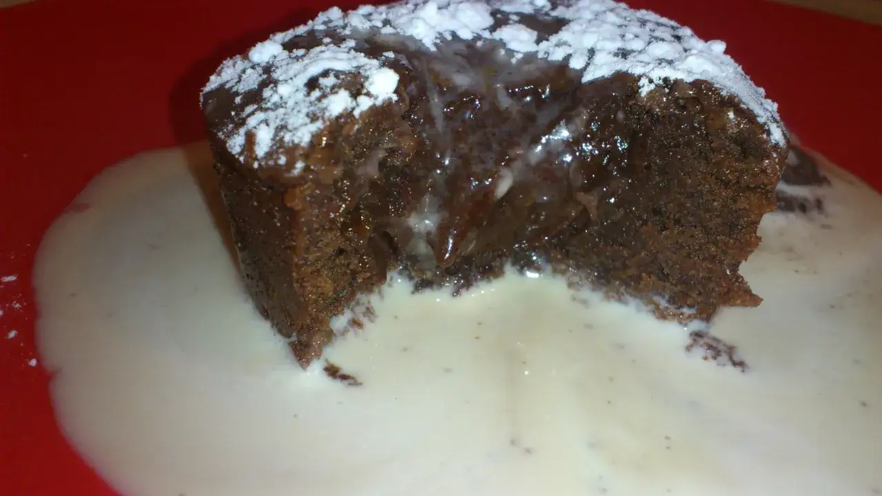 Un fondant au chocolat dégoulinant, saupoudré de sucre glace, servi dans une sauce crémeuse sur une assiette rouge.