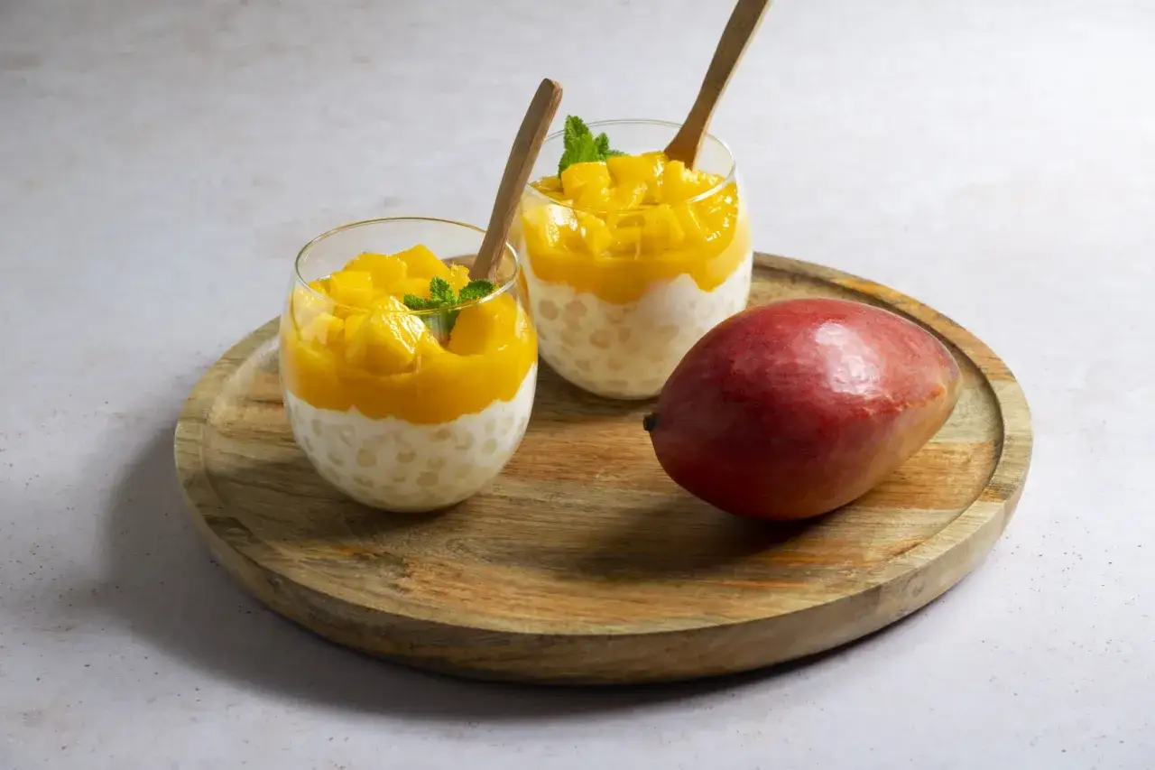 Pyszny deser z tapioki i mango, idealny sposób, by wykorzystać mrożone mango. Podany w szklankach z drewnianymi łyżeczkami.