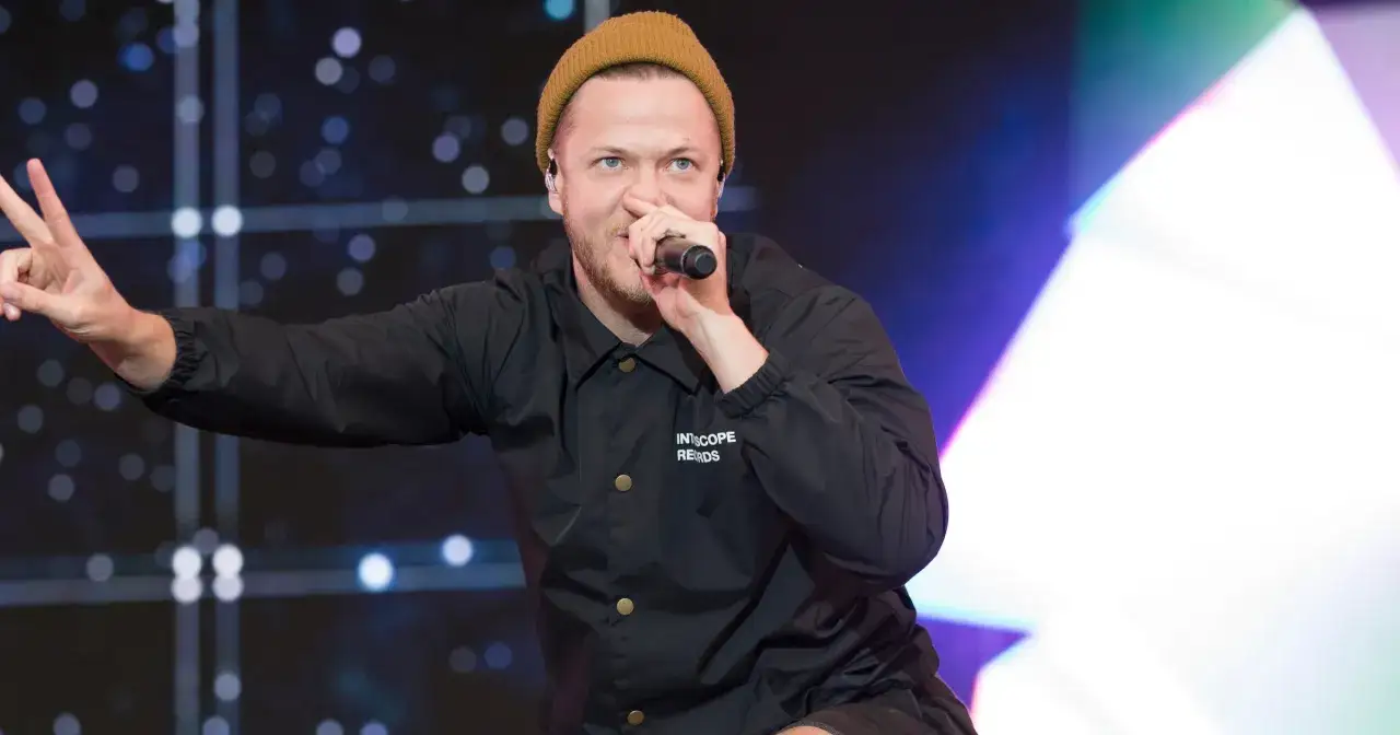 Imagine Dragons wokalista, Dan Reynolds, w beżowej czapce i czarnej kurtce, śpiewa do mikrofonu, pokazując znak pokoju.