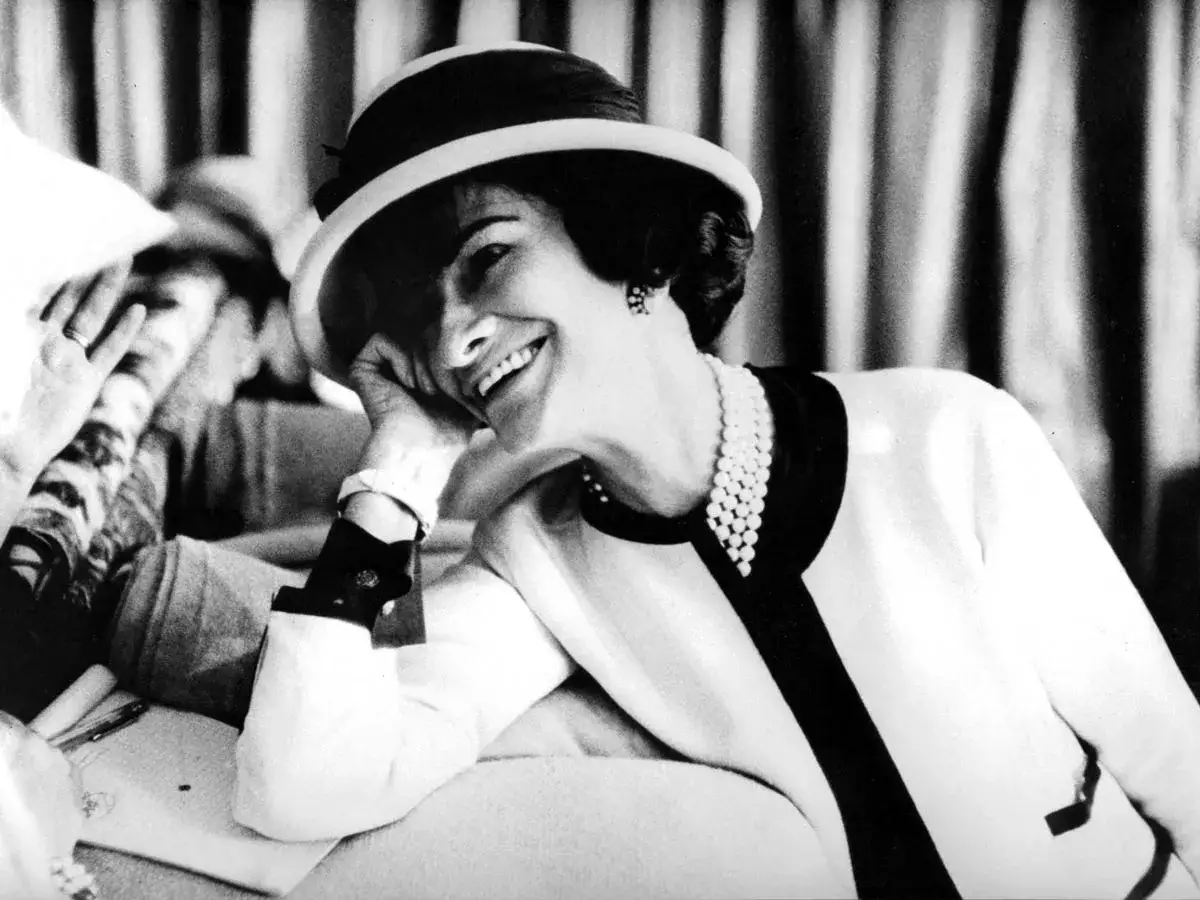 Coco Chanel, sonriendo con un sombrero y perlas, evoca la elegancia de donde es Chanel.