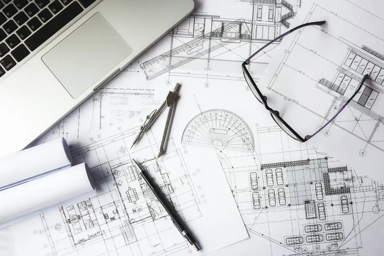 Na stole architekta leżą plany budynku, laptop, okulary i cyrkiel. To miejsce pracy nad projektem i jego kosztorysem.