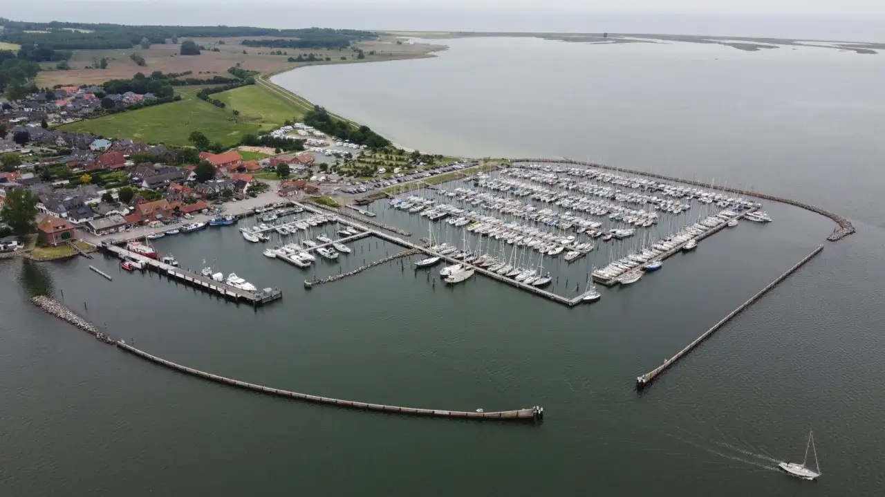 Maasholm Hafen mit vielen Segelbooten und Yachten, die an Stegen liegen. Im Hintergrund ein Dorf und das weite Wasser.