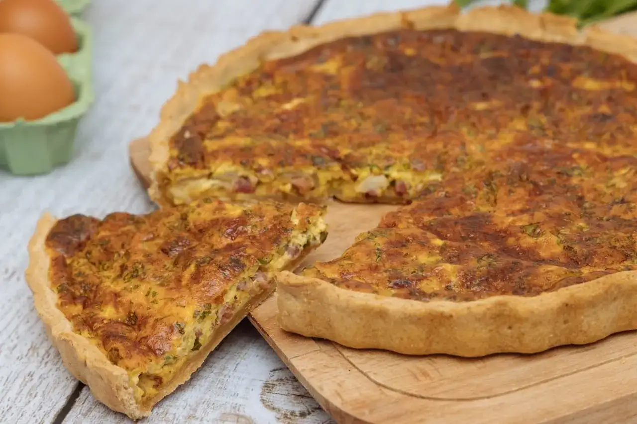 Une part de quiche lorraine, plat originaire de Lorraine, dorée et appétissante, sur une planche en bois. Des œufs frais sont visibles en arrière-plan.