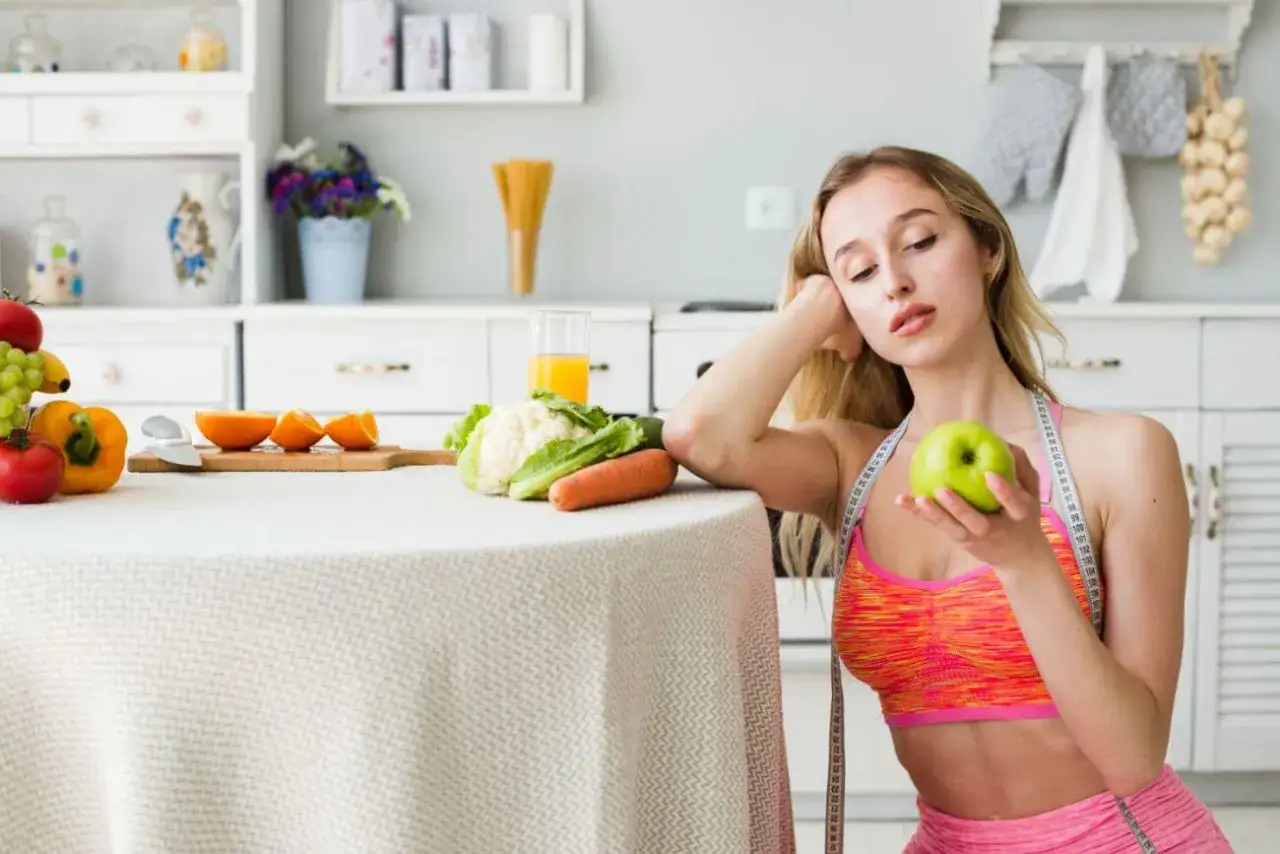 Młoda kobieta z miarką krawiecką na szyi trzyma jabłko, rozważając dietę 1100 kcal. Na stole warzywa i owoce.