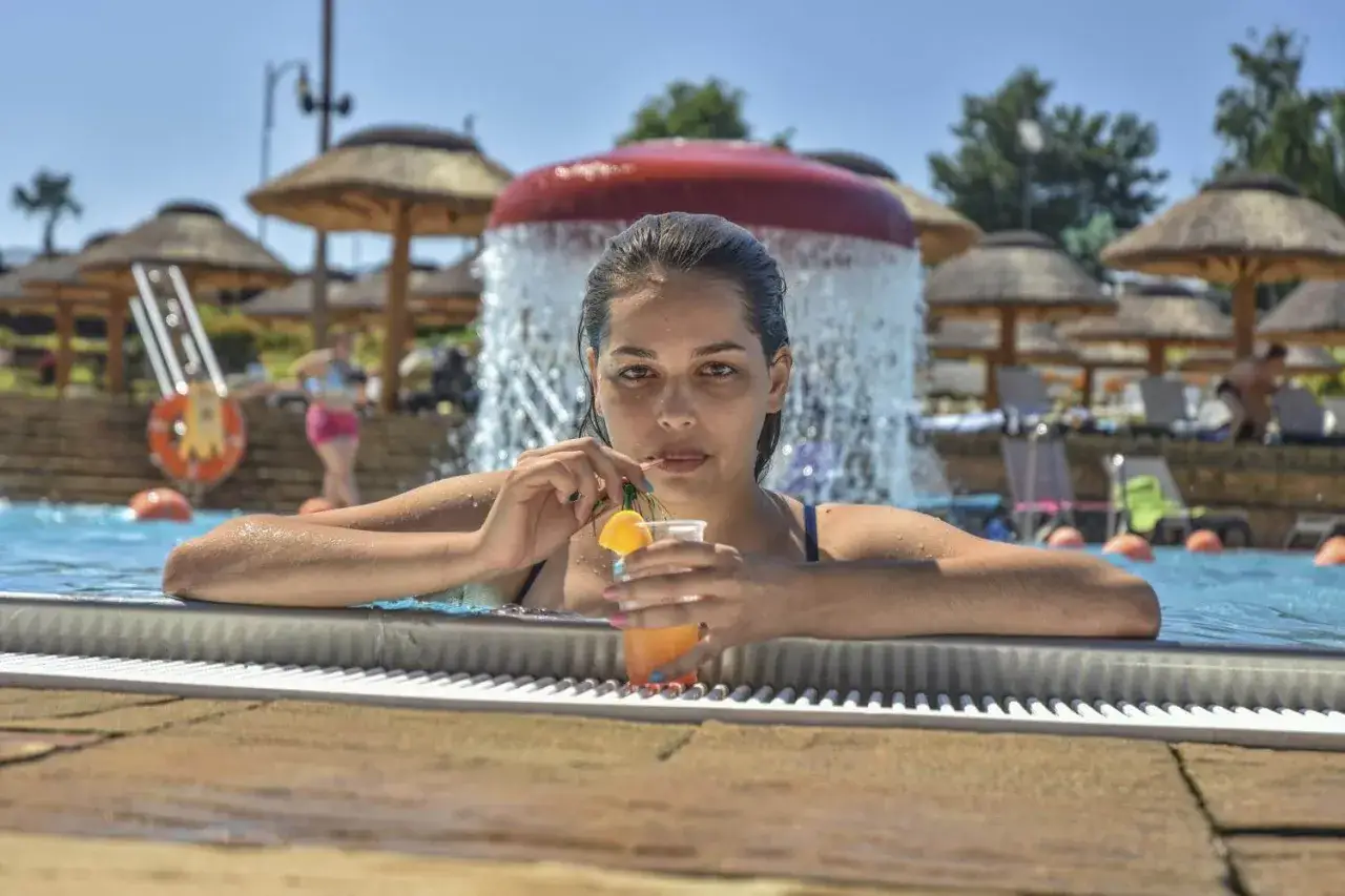Kobieta z drinkiem w basenie, otoczona palmowymi parasolami i wodnym placem zabaw. To właśnie co oznacza all inclusive – beztroski relaks.