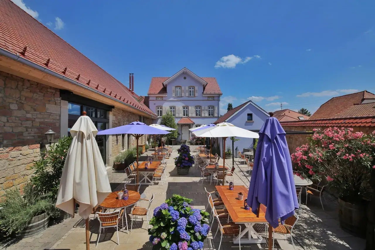 Terrasse eines Weinguts mit Tischen, Stühlen, Sonnenschirmen und Blumen unter blauem Himmel.