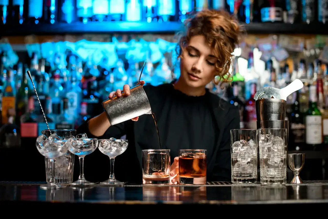 Barman nalewa drink do szklanki na tle butelek i niebieskiego światła.