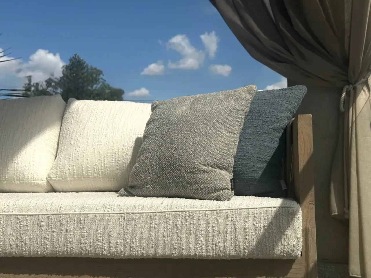 Wygodna sofa z poduszkami, osłonięta zasłonami, stoi na tle błękitnego nieba. Jej rama wykonana jest z pięknego, naturalnego **drewna tekowego**.