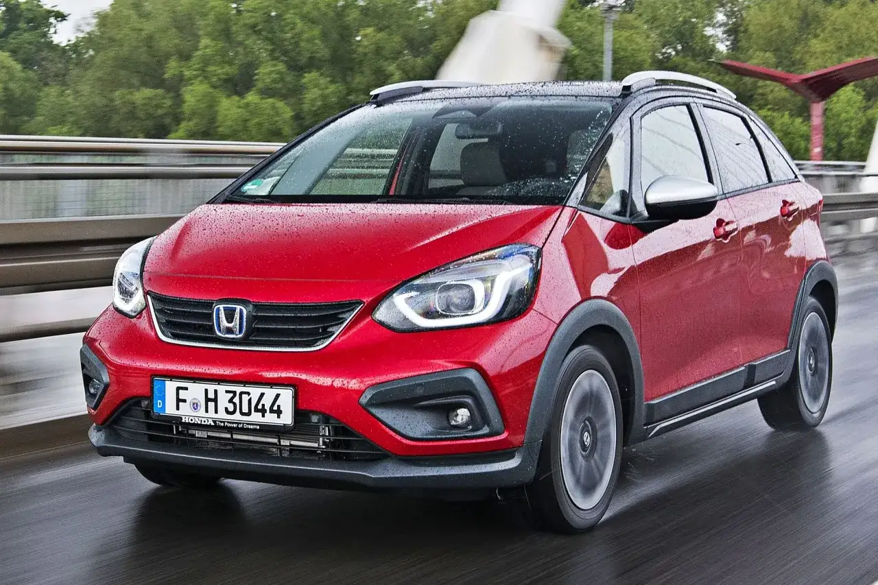 Czerwony Honda Fit w wersji Cross, z kroplami deszczu na karoserii, jedzie po mokrej drodze.