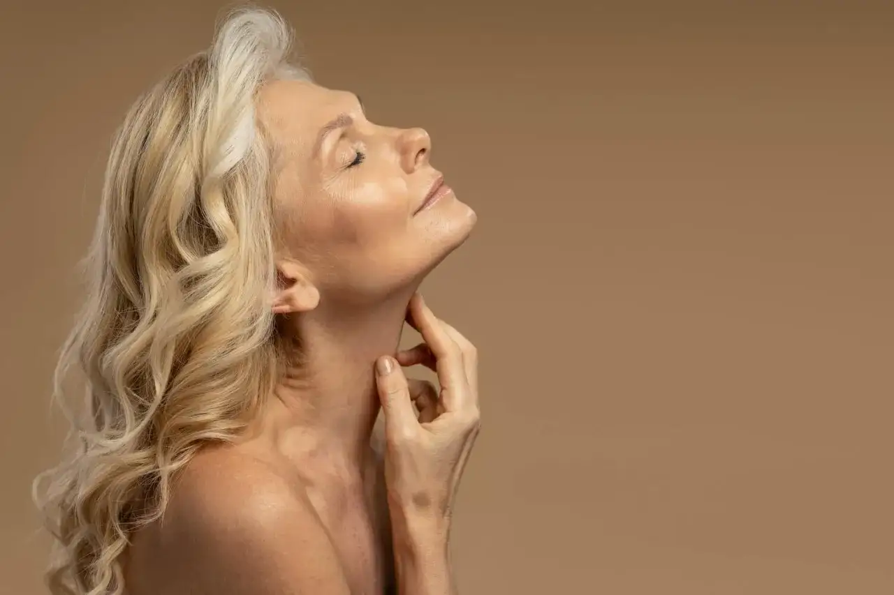 Kobieta z blond włosami i zamkniętymi oczami delikatnie dotyka szyi. Jej skóra w okresie menopauzy jest widoczna, podkreślając naturalne piękno.