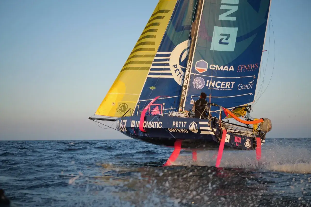 Segelboot mit pinken Foils rast über die Wellen. Ein Skipper steuert das Boot bei der Mini Transat Regatta.