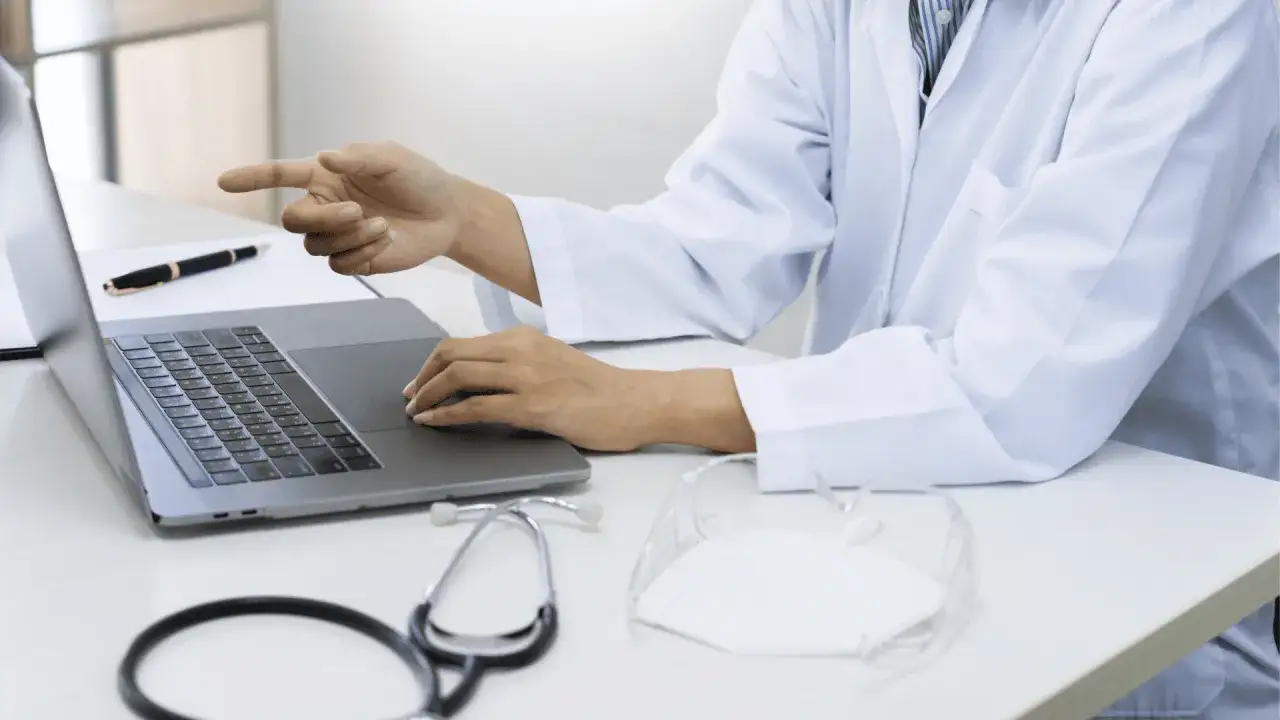 Lekarz w białym kitlu przy laptopie wyjaśnia, czy do poradni dermatologicznej potrzebne jest skierowanie. Na biurku stetoskop i maseczka.