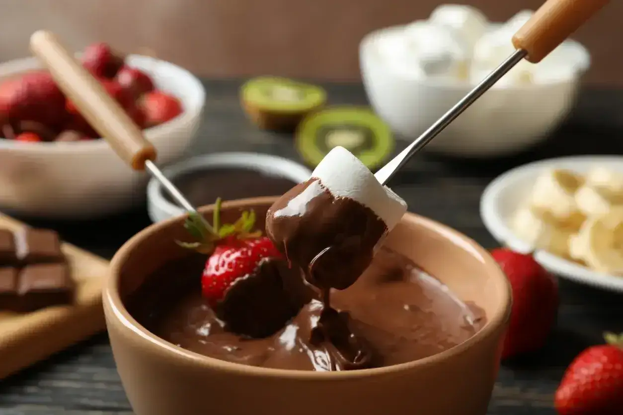Pyszne fondue czekoladowe z pianką i truskawką na patyczku. W tle owoce i pianki.