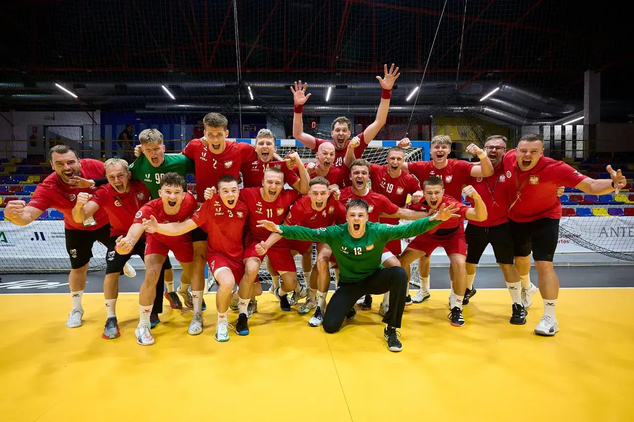 Drużyna piłkarzy ręcznych w czerwonych strojach świętuje zwycięstwo. Wśród nich Krzysztof Zimny, bramkarz w zielonym stroju, klęczy z wyciągniętymi rękami.