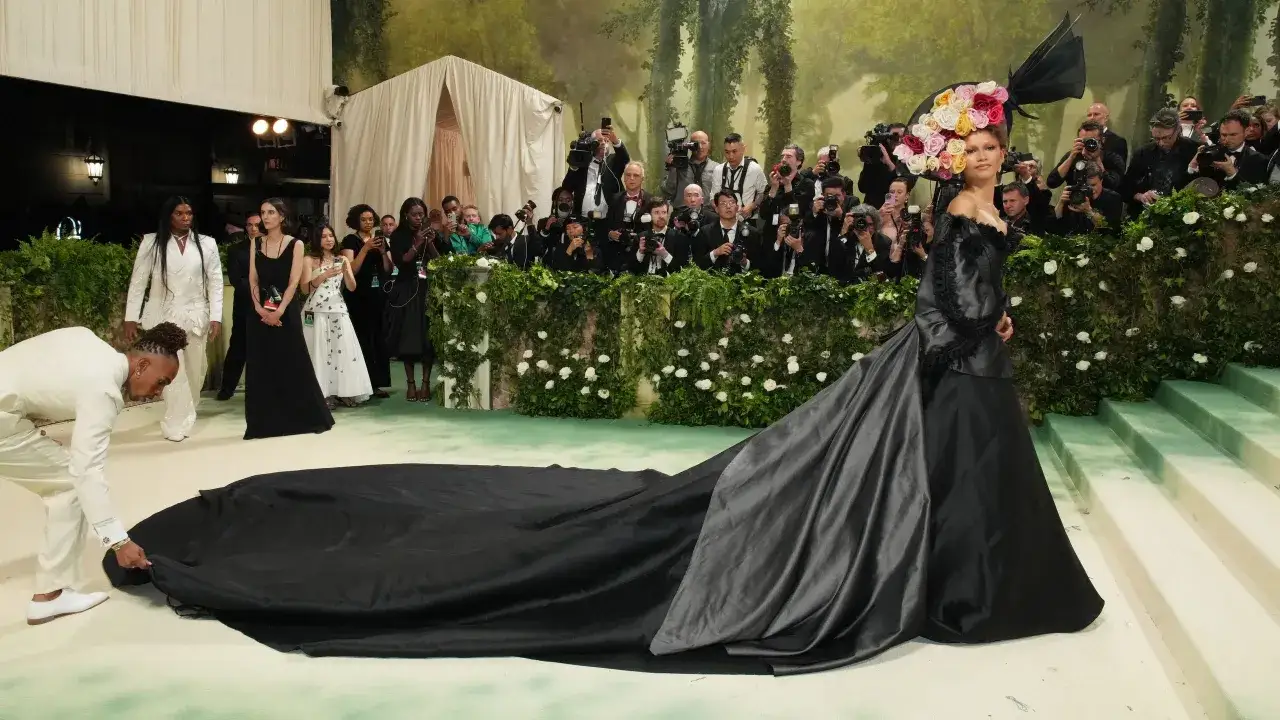 Una mujer con un vestido negro y un tocado floral posa para las cámaras en la alfombra verde, mientras un hombre ajusta la cola de su vestido, todo bajo el tema de la Met Gala.