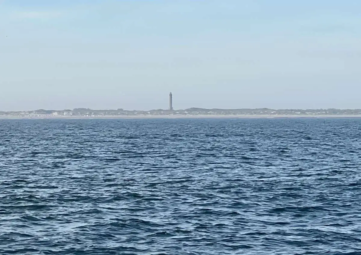 Der Leuchtturm Norderney ragt über die Dünenlandschaft am Horizont. Das weite Meer liegt davor.