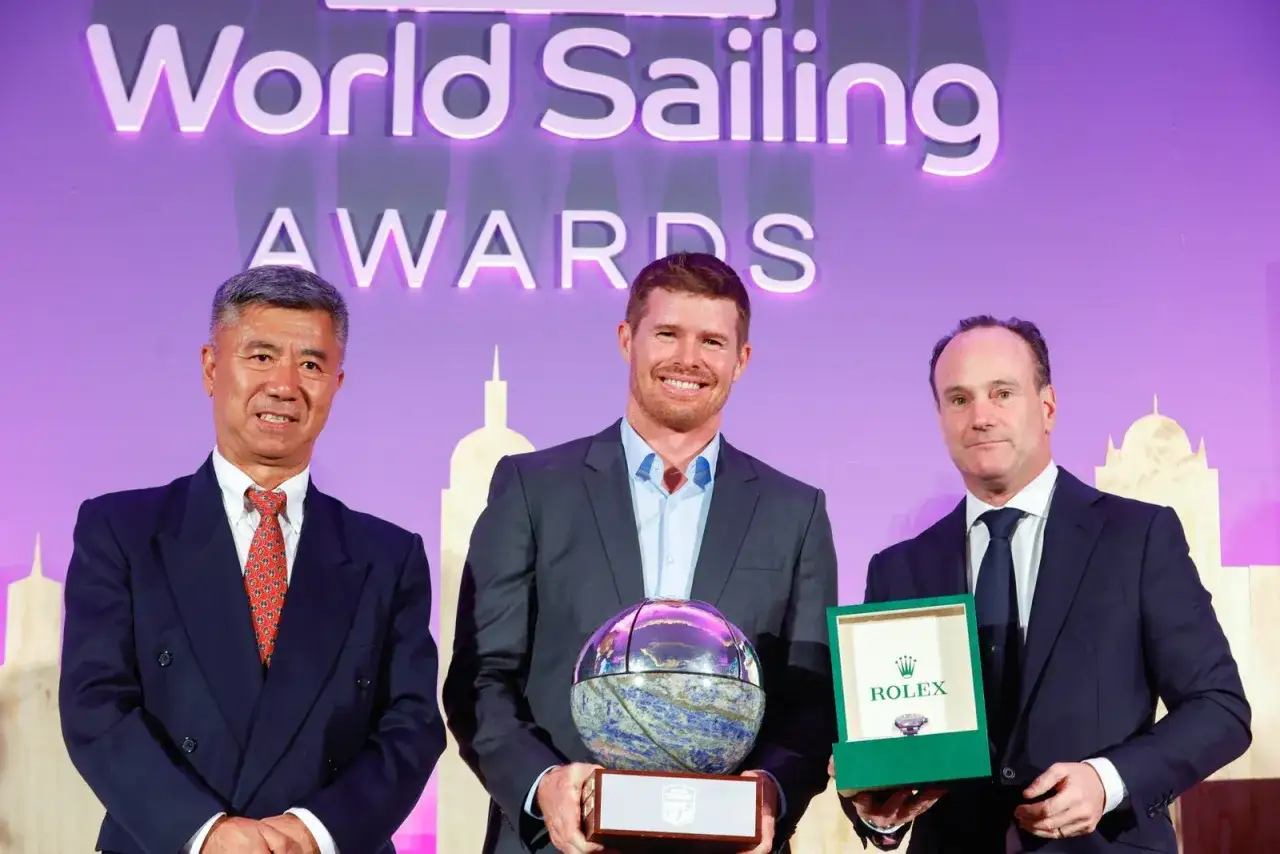 Tom Slingsby mit Auszeichnung bei den World Sailing Awards. Er hält eine Trophäe, daneben zwei Männer mit Geschenken.