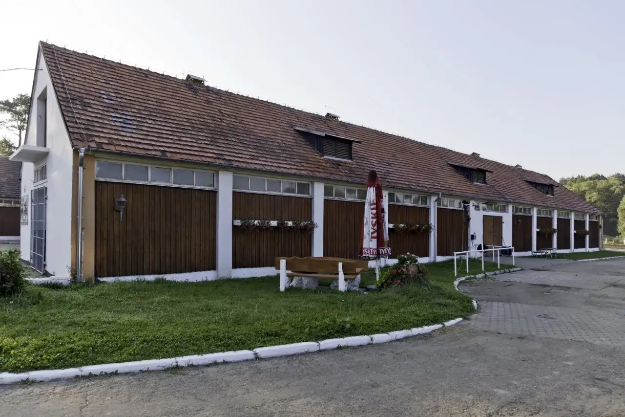 Długi budynek stajni z drewnianymi drzwiami i oknami, z parasolem i ławką przed nim.