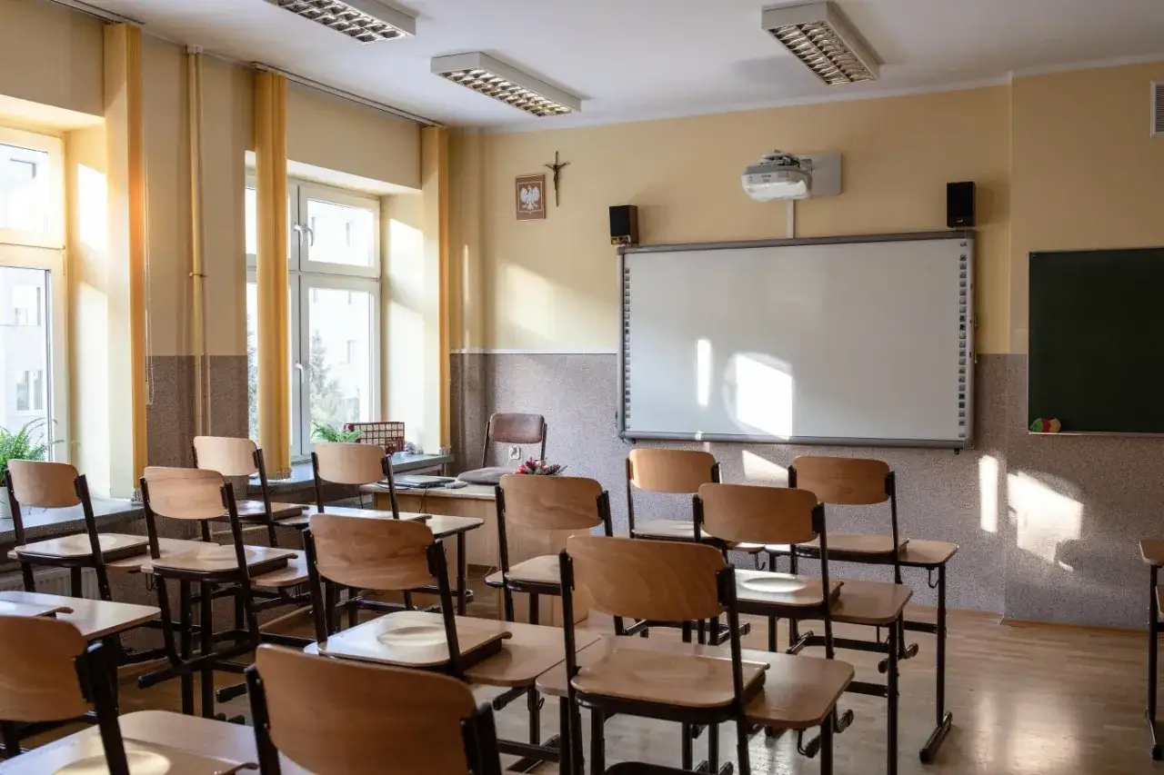 Pusta klasa z ławkami, tablicą interaktywną i krzyżem. Czy religia wlicza się do czerwonego paska? Słońce wpada przez okna.